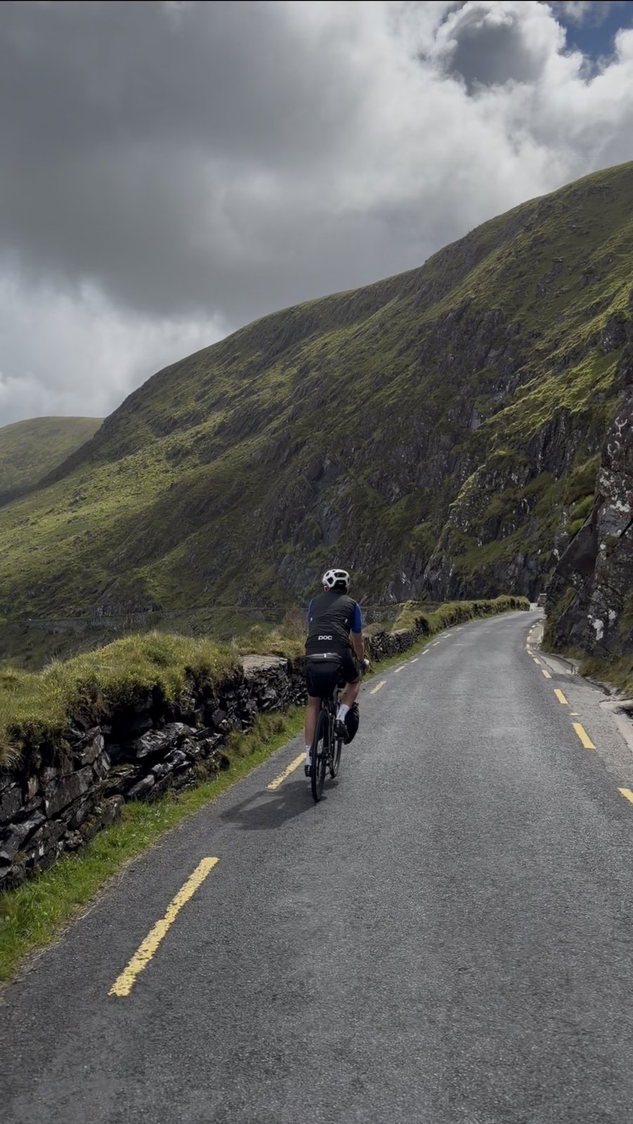 A rider climbing the Conor Pass with a steep green mountain wall rising above the narrow road