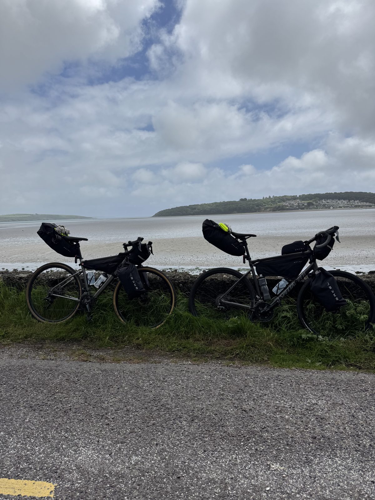 Two loaded bikepacking bikes leaning against a low wall beside a tidal inlet at low tide