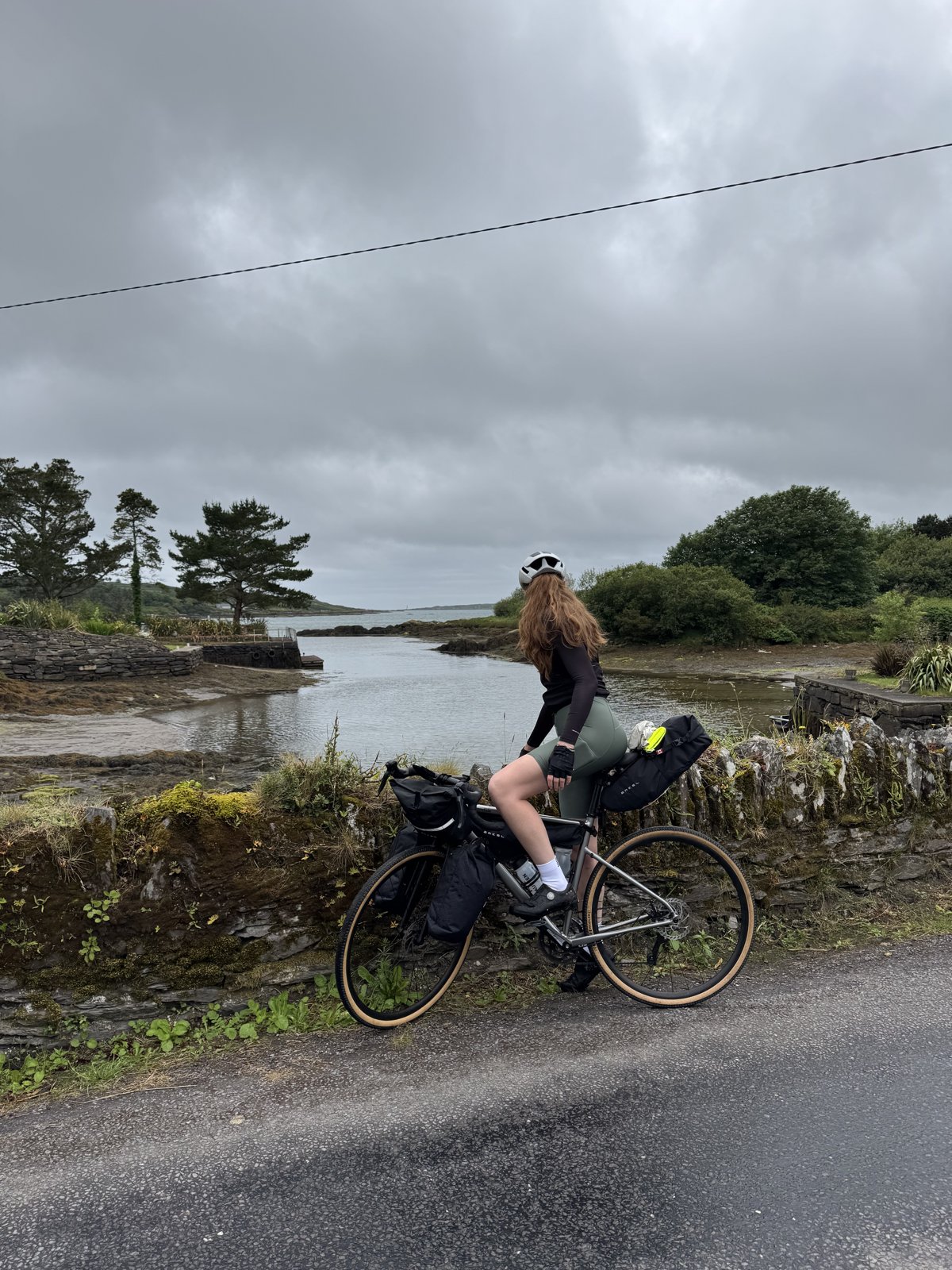 Bailey riding the loaded bike past a tidal inlet on the Mizen under a dramatic grey sky