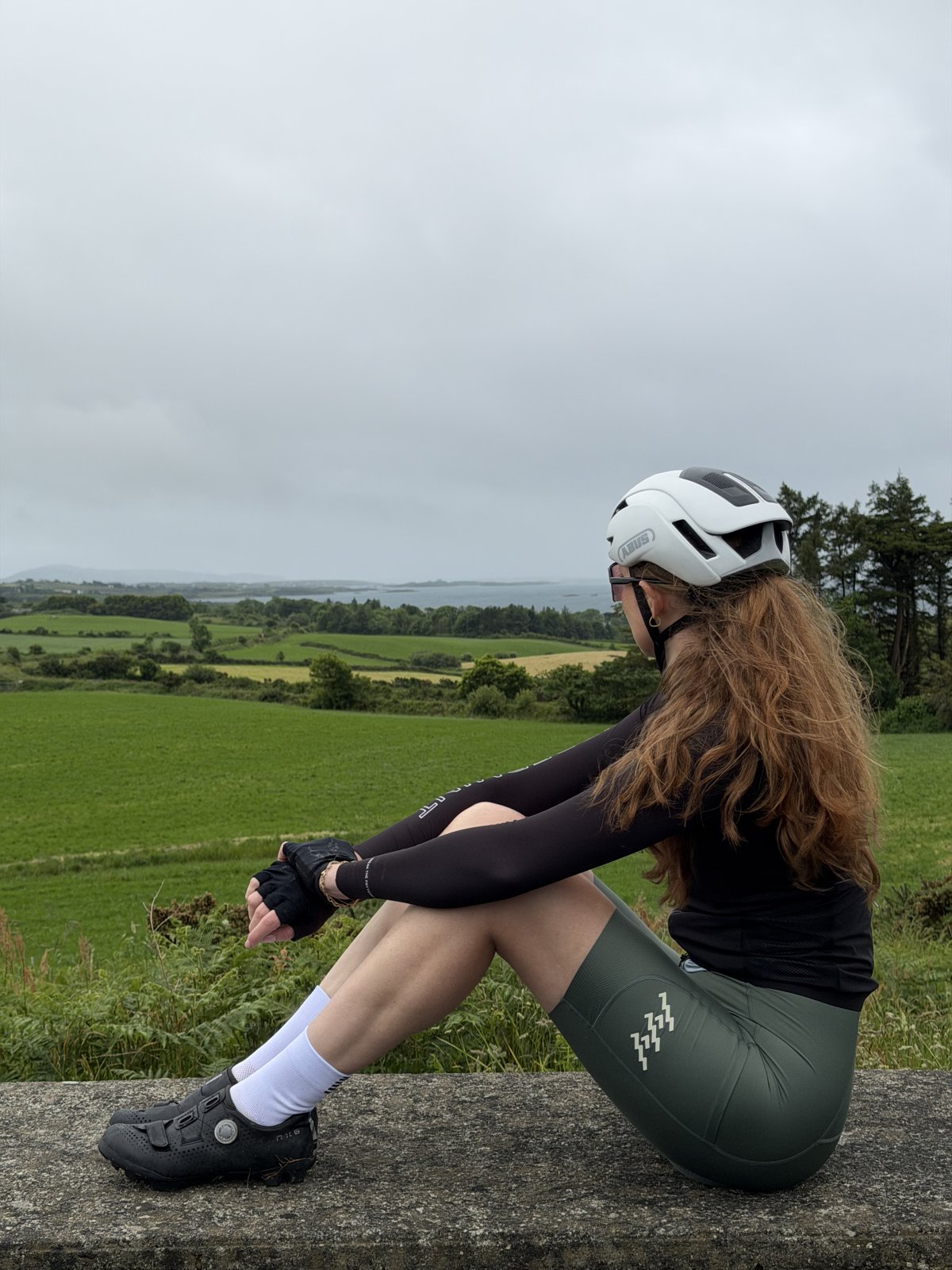 Bailey resting on a wall in cycling kit, looking out over green fields to a West Cork inlet