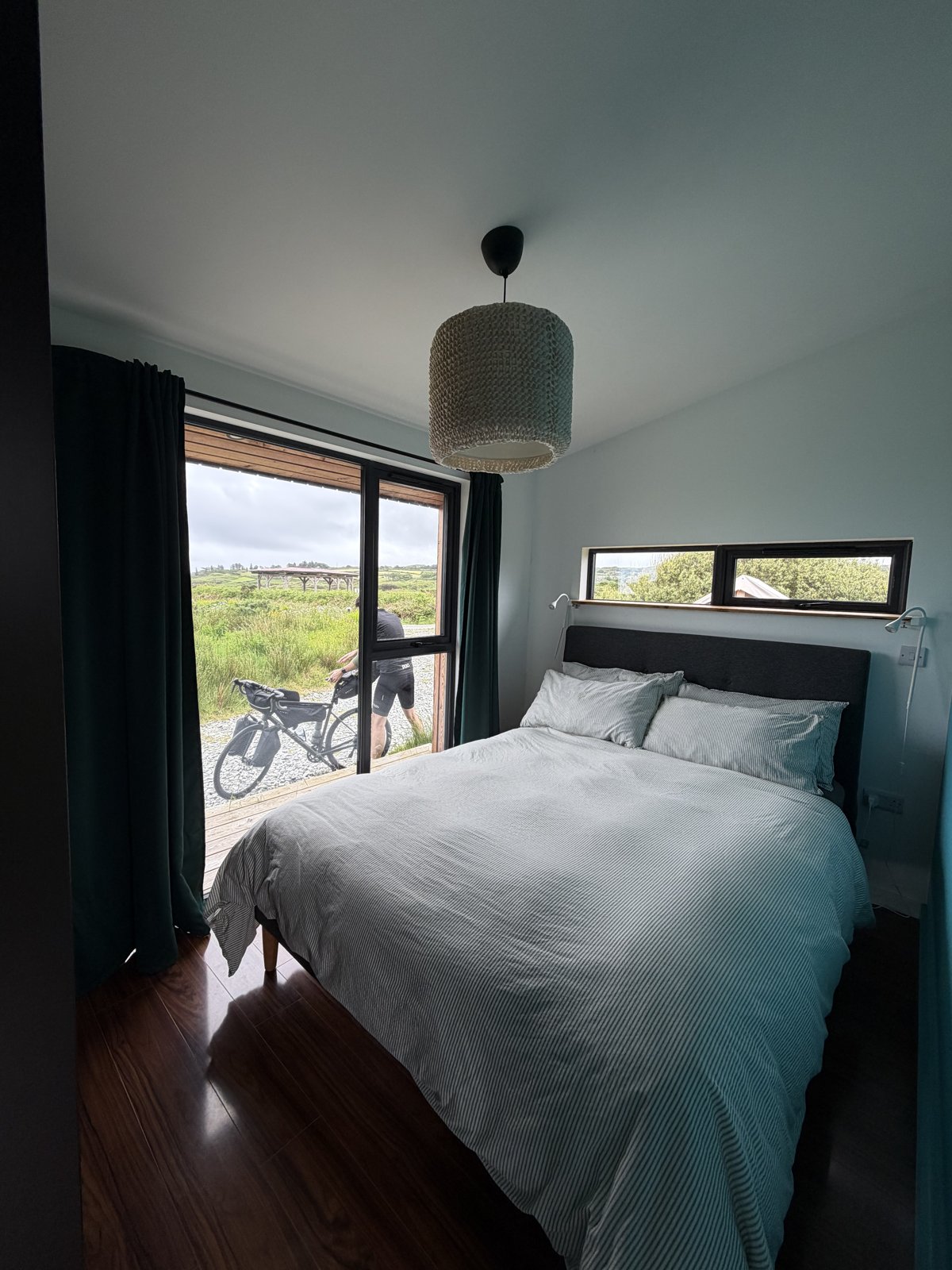 The cabin bedroom with a big window looking out over the fields and a bike parked on the deck