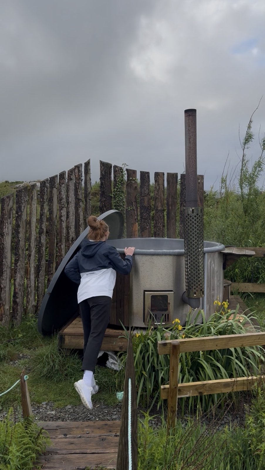 Bailey climbing into the wood-fired hot tub at Goleen Harbour, behind a timber fence under a grey West Cork sky