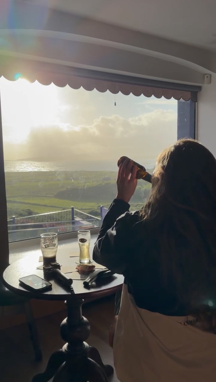 Bailey raising a pint at the window of Causkey's Bar, Coulagh Bay and the evening sun spread out below