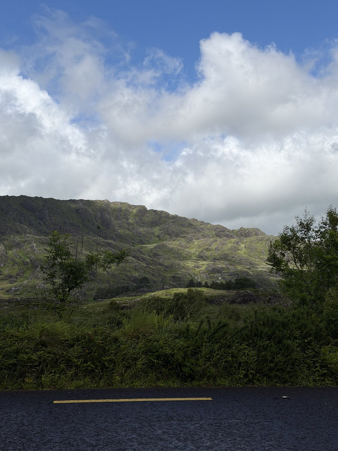A rugged green mountainside rising above the road on the Beara, clouds breaking to blue overhead