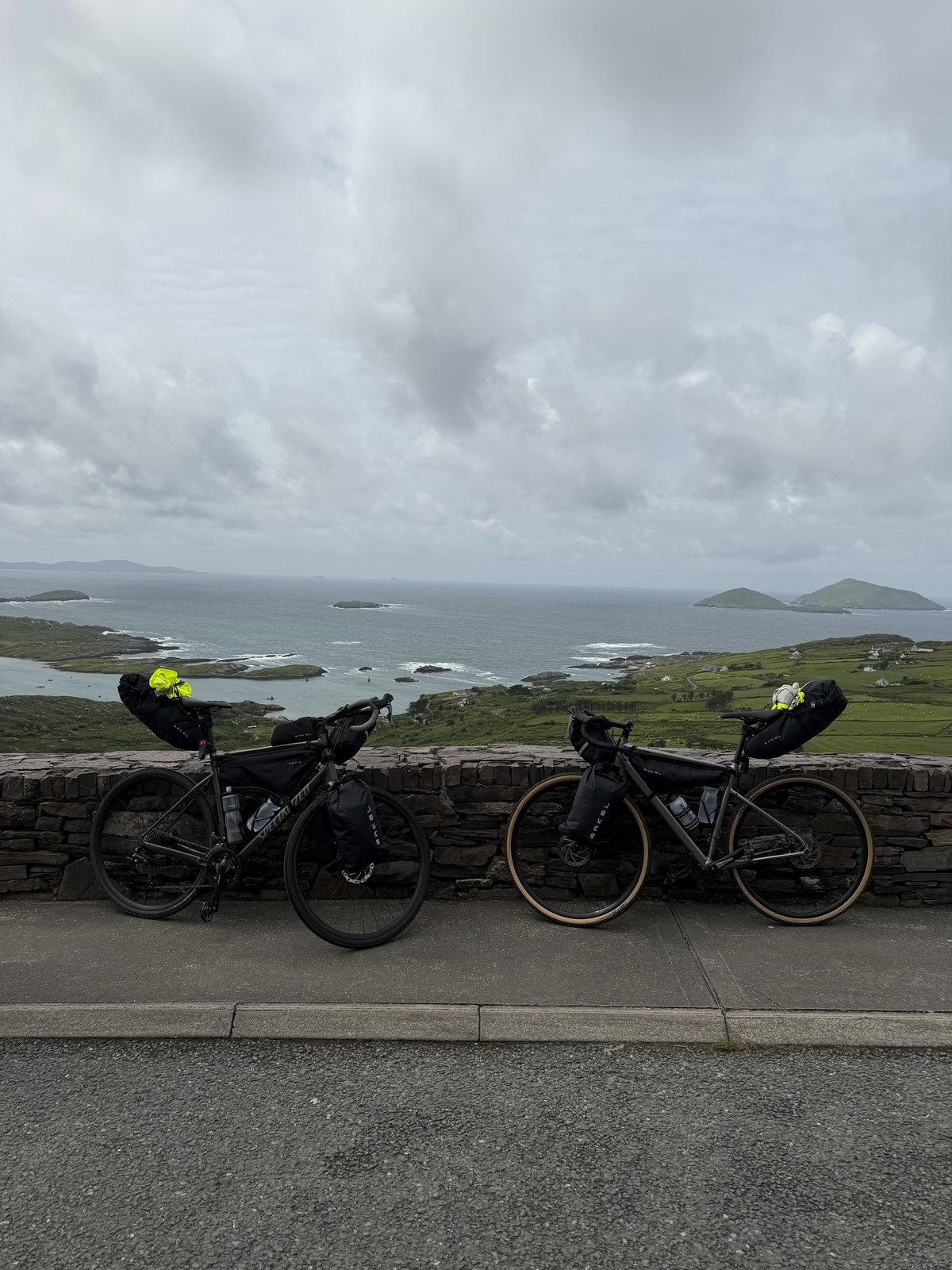 Two loaded bikes leaning on a stone wall above a wide Ring of Kerry bay dotted with small islands under a grey sky