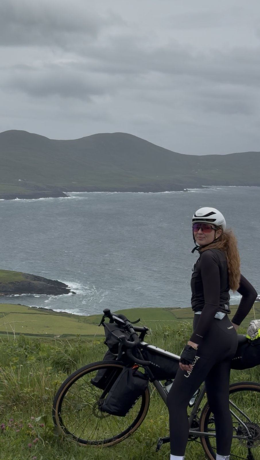 Bailey astride her loaded bike, looking back over a bay and green fields toward a mountain on a moody day