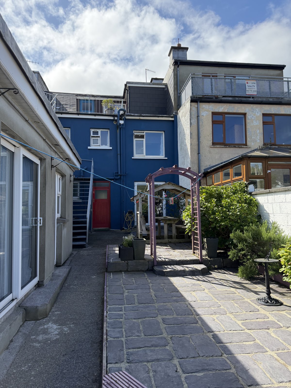 The blue-painted rear courtyard of Síve Hostel in Cahersiveen, with a red door, a garden arbour and paved patio in the sun