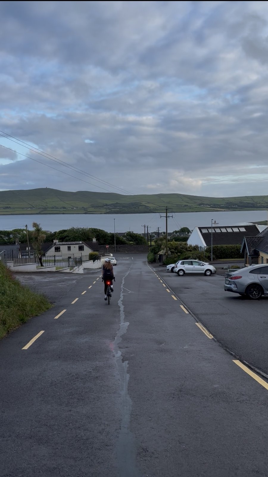 A rider freewheeling down a road toward Ventry harbour with the hills of the Dingle Peninsula across the bay
